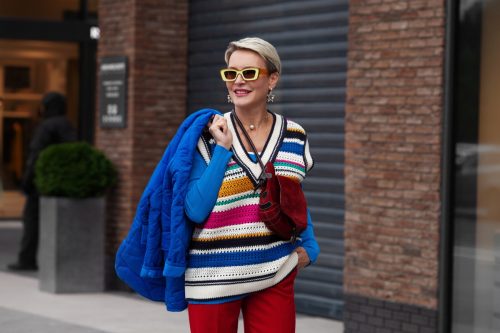 Smiling Happy Model is wearing blue jacket and sweater, striped gilet, red pants and yellow sunglasses. Fashion Street Style autumn or spring. Woman posing outside the clothing store