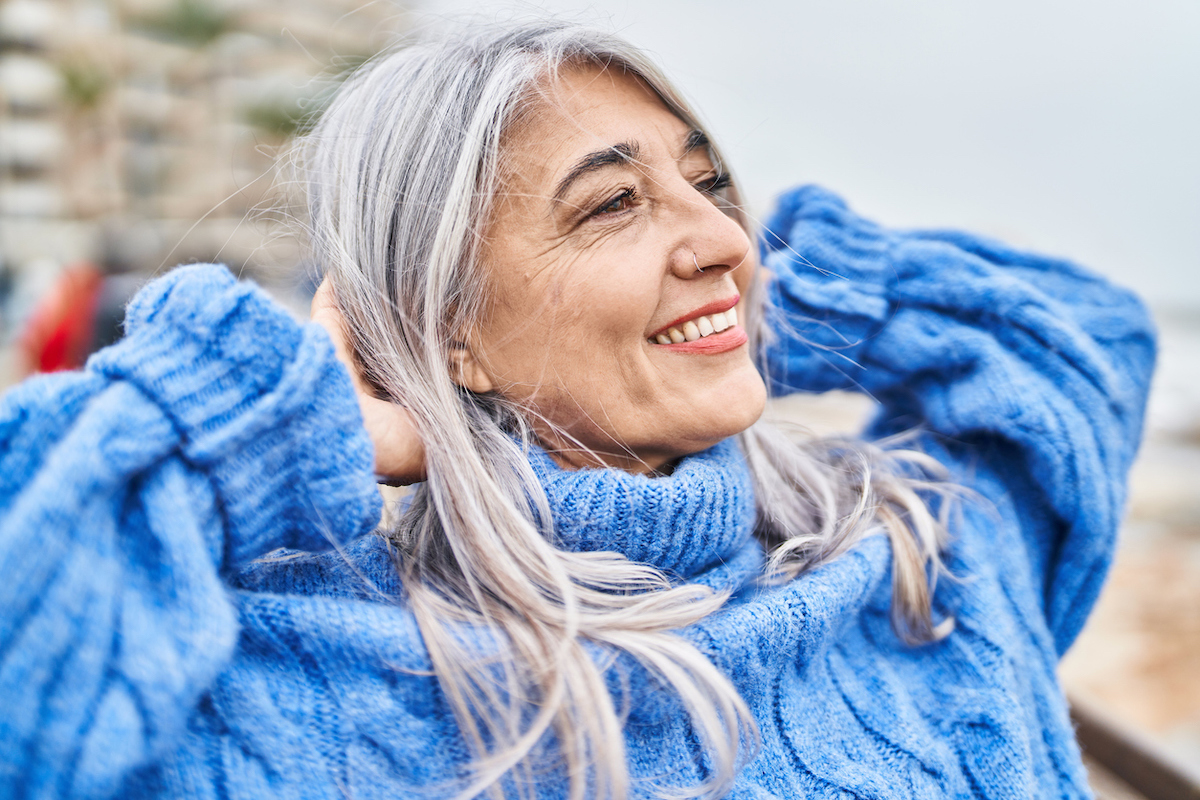 Middle age gray-haired woman smiling with hands on head at seaside, wearing a blue sweater