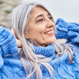 Middle age gray-haired woman smiling with hands on head at seaside, wearing a blue sweater