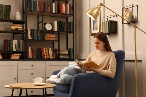 Young woman reading book in armchair in home library.