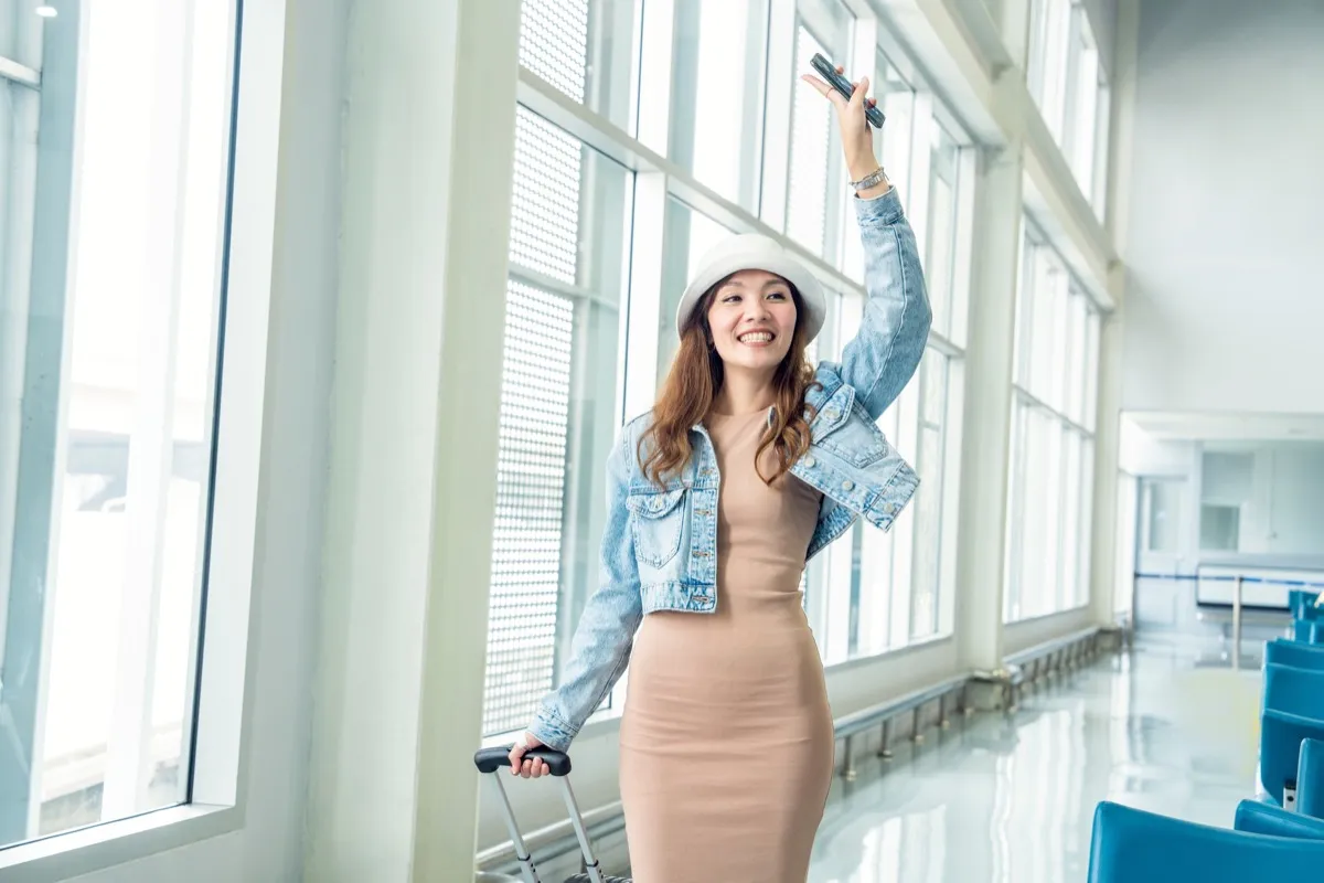 Asian female tourist backpacking in denim jacket dress dragging luggage, raising hands, cheering and greeting friends in airport terminal
