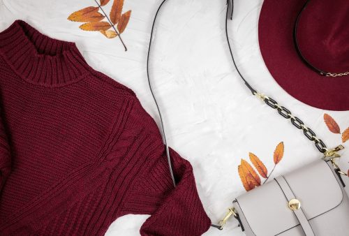 Flat lay of a woman's burgundy sweater, burgundy fedora, and taupe leather purse against a light wood background.
