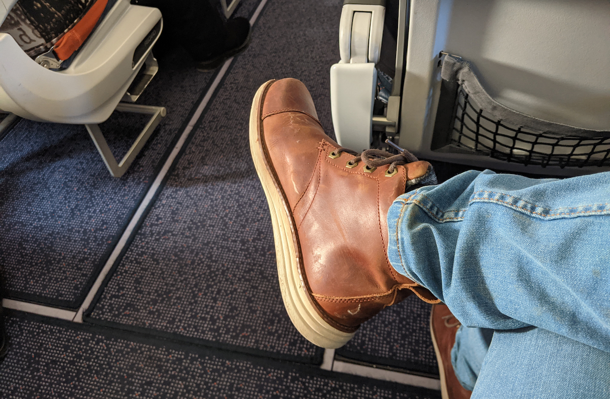 Close up of a man stretching his legs into the aisle while seated on an airplane, wearing jeans and brown leather boots