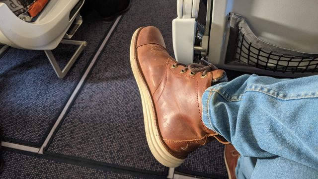 Close up of a man stretching his legs into the aisle while seated on an airplane, wearing jeans and brown leather boots