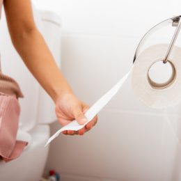 Close up of a woman wearing a light pink dress sitting on a toilet pulling toilet paper