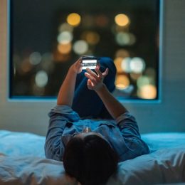 A woman is lying down on a bed and using a smart phone at night.