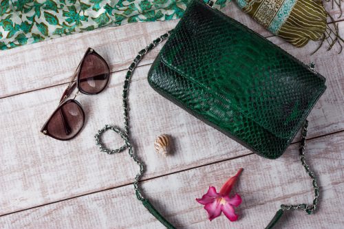 Flat lay of an emerald green snakeskin purse, green patterned scar, and sunglasses on a light wood background.