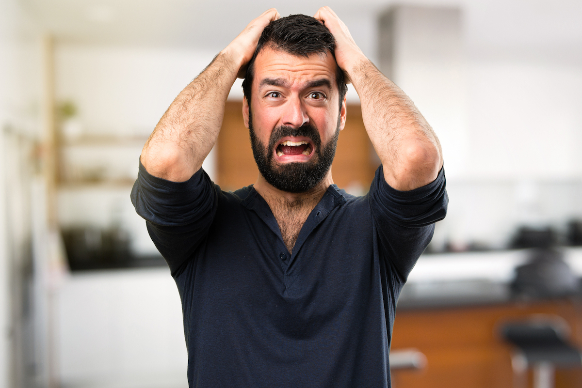A frustrated young man with a beard inside his home grabbing his head and yelling.