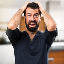 A frustrated young man with a beard inside his home grabbing his head and yelling.