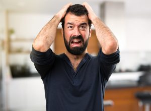 A frustrated young man with a beard inside his home grabbing his head and yelling.