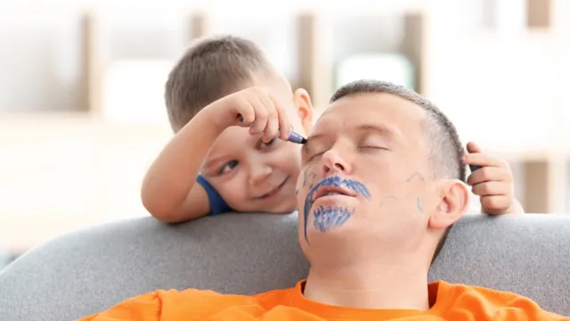 little boy drawing on his father's face as he sleeps
