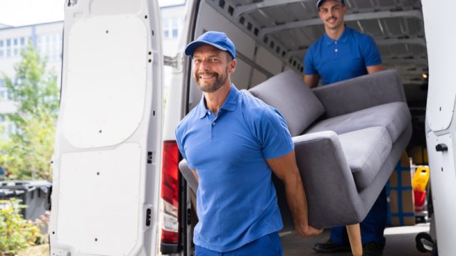 two delivery men taking a couch out of their truck and smiling at the camera