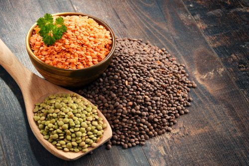Green, black, and red lentils in piles on a wood table.