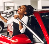 A happy young woman getting into a new red car in a car dealership.