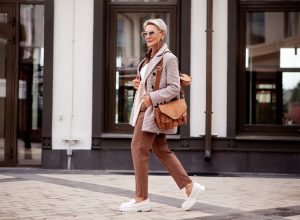 Side view full length fashionable woman walks outside and wears trendy clothes, oversize jacket, pants, stylish bag and white loafer shoes. Confident female model with short blonde hair