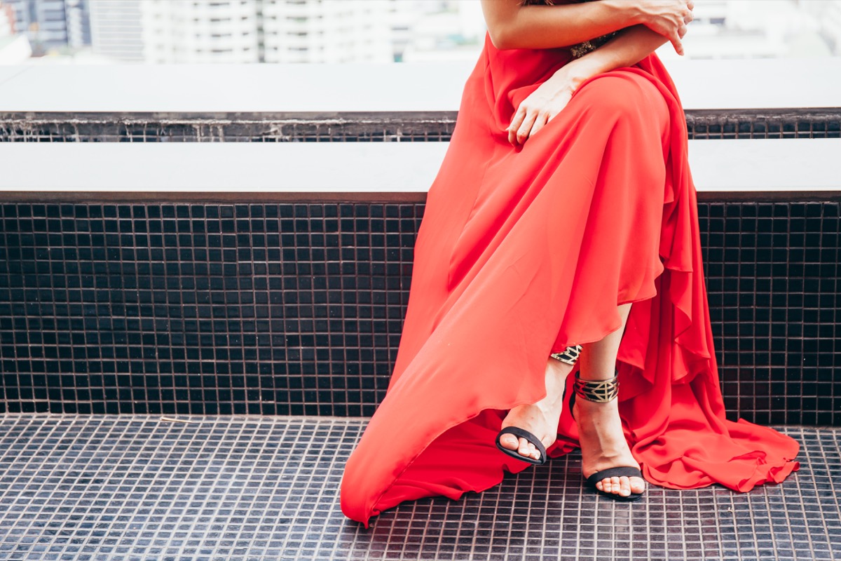 Fashionable woman sitting crossed legs in long red dress in urban scene behind