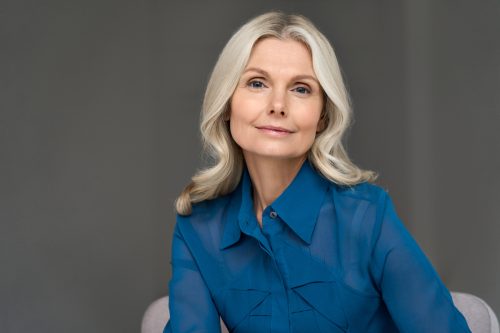 Sophisticated, elegant mature blonde woman wearing a blue blouse posing for a photo against a gray background.