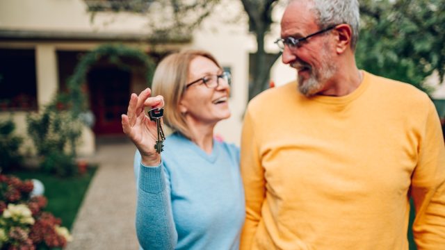 Senior couple smiling and holding up new house key while standing outside.