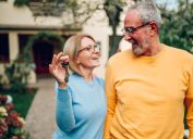 Senior couple smiling and holding up new house key while standing outside.
