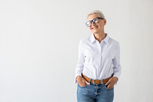 A mature woman with short white hair is wearing a white button-down shirt with her hands in her jeans pockets against a white background.