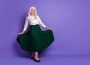 A happy mature woman with long white hair wearing a white silk blouse and long emerald green skirt against a purple background
