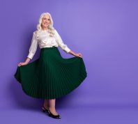 A happy mature woman with long white hair wearing a white silk blouse and long emerald green skirt against a purple background