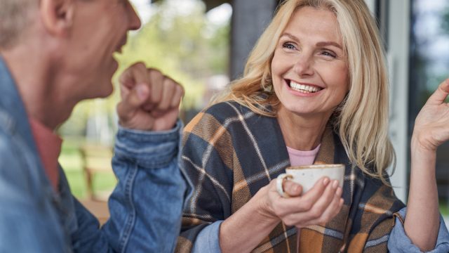 Cheerful mature lady is communicating with husband while relaxing with mugs of tea in green countryside
