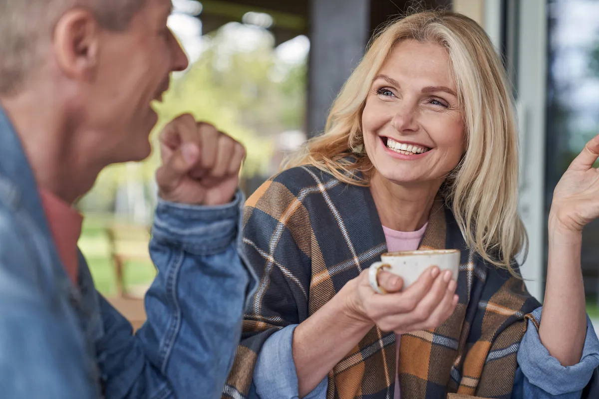 Cheerful mature lady is communicating with husband while relaxing with mugs of tea in green countryside