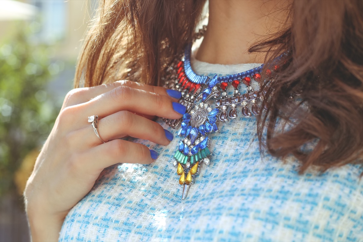 Brown-haired girl in a blue dress with a necklace. Large necklace rhinestones. Thin hand with blue nail. Fashion trends. Street fashion. Trendy, stylish. Fashionable girl.