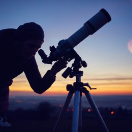 A person looking through a telescope at dawn or dusk with the moon and planets visible