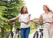 Two mature women in conversation while walking with bicycles at park.