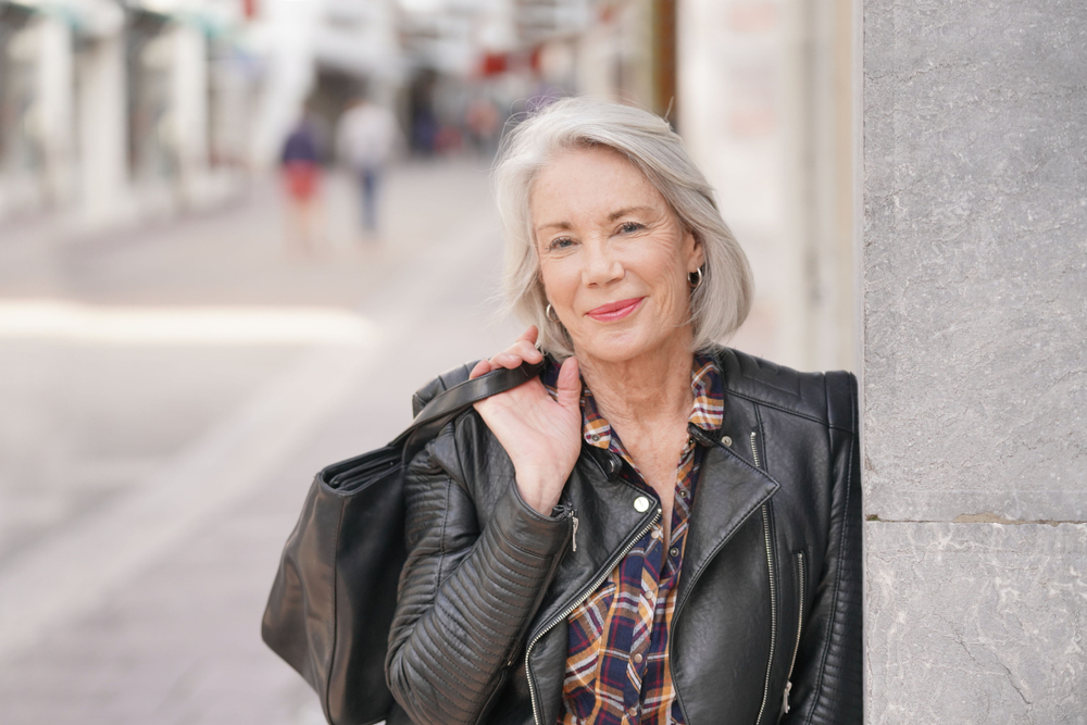 Stylish senior woman in black leather jacket leaning against a wall on a city street