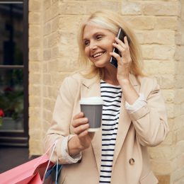 Stylish older woman smiling on phone with trench coat, holding a shopping bag and coffee