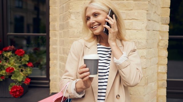 Stylish older senior woman smiling on phone with trench coat, shopping bag and coffee
