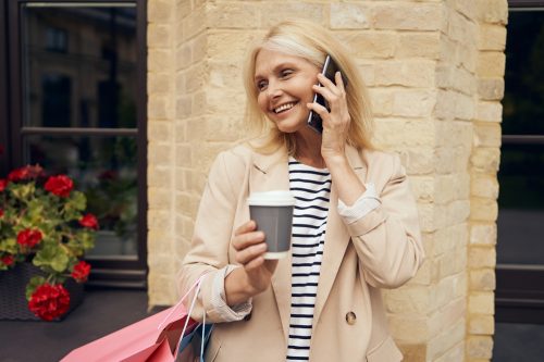 Stylish older woman smiling on phone with trench coat, holding a shopping bag and coffee
