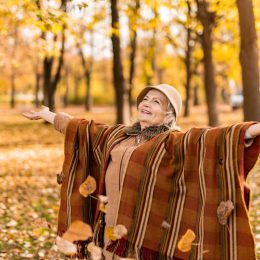 Happy senior woman wearing a poncho enjoying an autumn day in the park and throwing leaves.