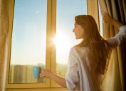 A smiling young woman with long brown hair holding a cup of coffee and opening the curtains on a sunny morning.