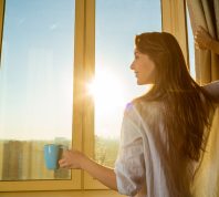 A smiling young woman with long brown hair holding a cup of coffee and opening the curtains on a sunny morning.