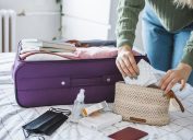 Woman preparing travel suitcase on bed at home. She is packing medicine and disinfection stuff for travel.