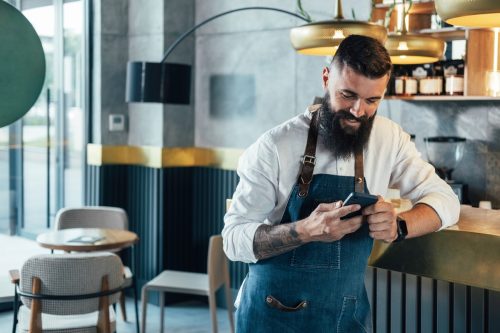 Cheerful smiling waiter with a beard leaning on the bar counter and typing text message on his smartphone while working at the coffee shop.