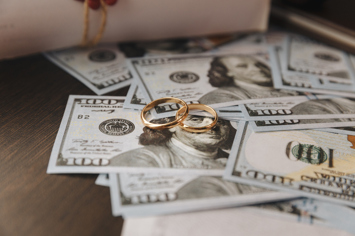 Two gold wedding bands sitting on top of a pile of hundred dollar bills.
