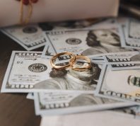 Two gold wedding bands sitting on top of a pile of hundred dollar bills.