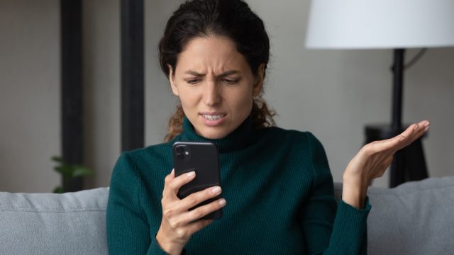 A young woman sits on her couch and looks at her phone annoyed.