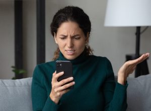 A young woman sits on her couch and looks at her phone annoyed.