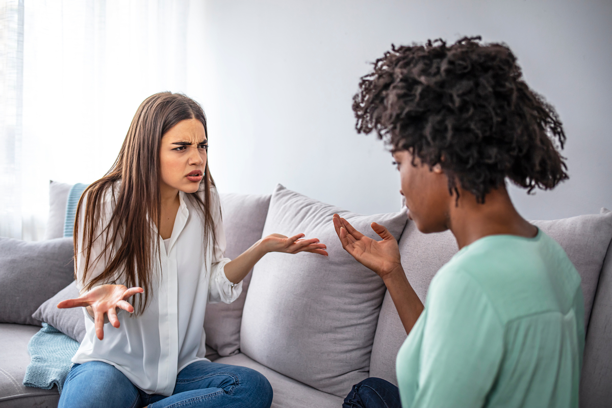 A young woman sitting on her couch looking offended by her female friend.