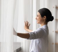A young brunette woman opens her sheer window curtains and smiles.