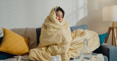 Sick young woman headache fever cough cold sneezing sitting under the blanket on sofa in living room at home.