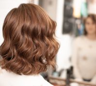 Back of a woman's head sitting at a mirror with shiny, wavy auburn hair