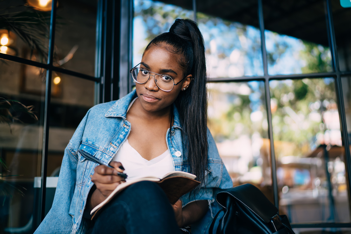 A young Black woman wearing a jean jacket and her hair in a ponytail sits outside and writes in a journal.