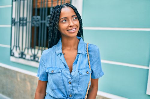 Young Black woman with braided hair and a denim shirt smiling while walking in the city.